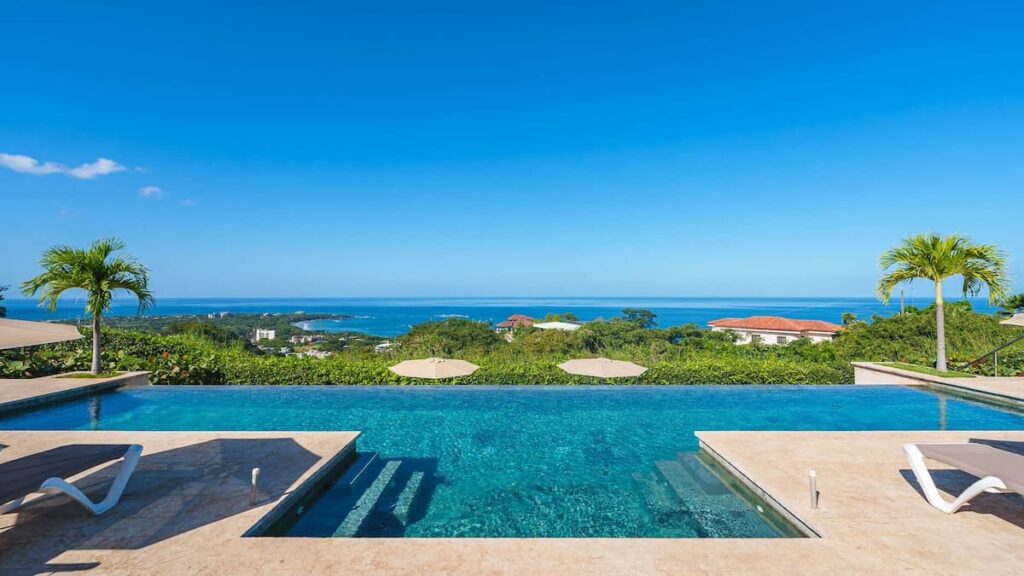 La Vista vacation rental's pool overlooking the Pacific Ocean on a sunny day with palm trees either side of the pool