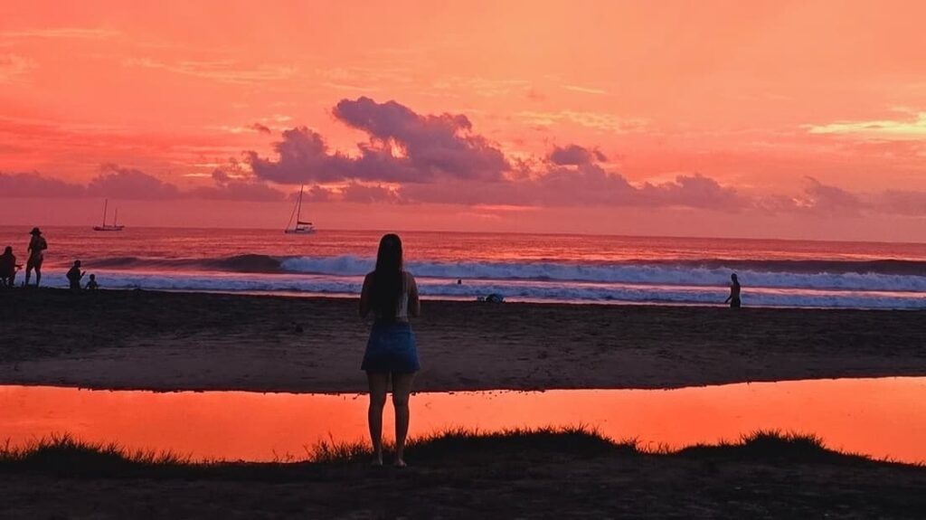 Woman looking at the pink sunset from the El Vaquero beach bar in Tamarindo with large sailboats in the distance