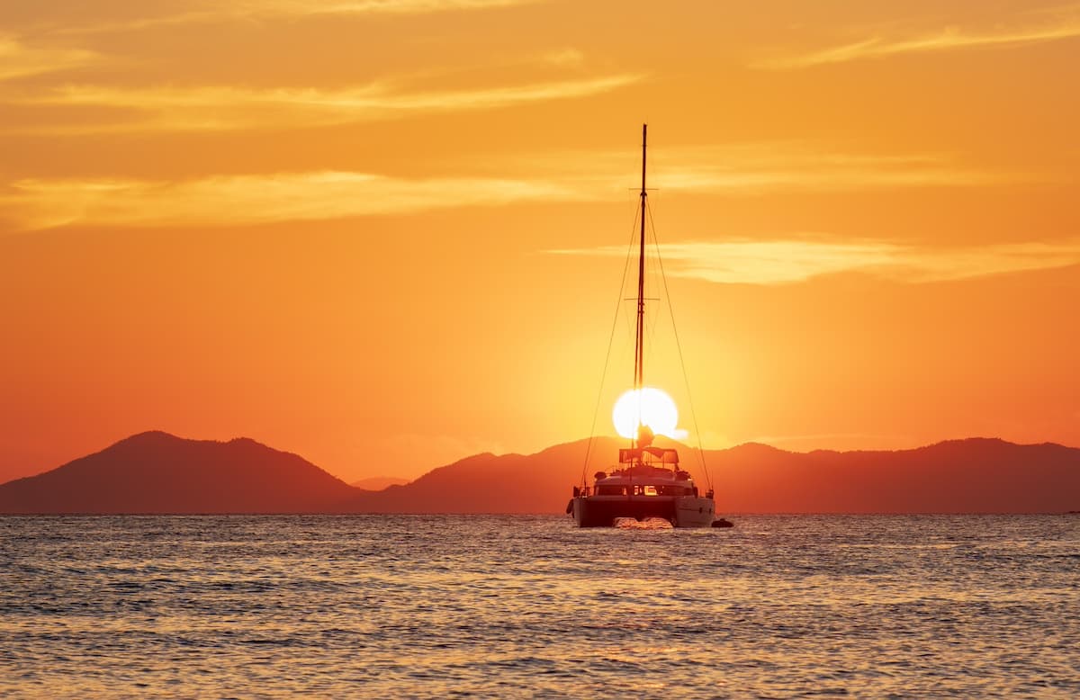 Large sailboat out at sea with large orange sun setting in the background behind mountains