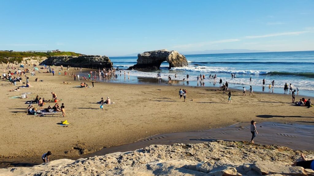 Crowds sunbathing and swimming on Naturl Bridges State Beach with natural bridge rock in background