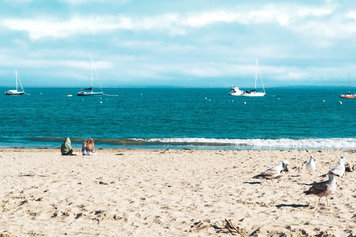 Two people sitting on sandy Capitola beach with seagulls and boats in the water