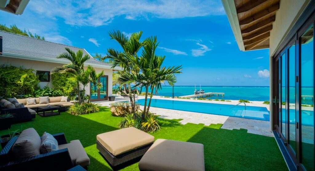Luxurious ocean front villa with a lush garden, infinity pool, and elegant outdoor lounge area, boasting a panoramic view of the clear blue ocean and a sky dotted with clouds