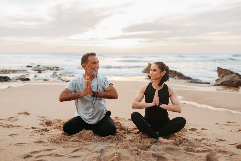 Man and woman doing yoga on a sandy beach with ocean in the background