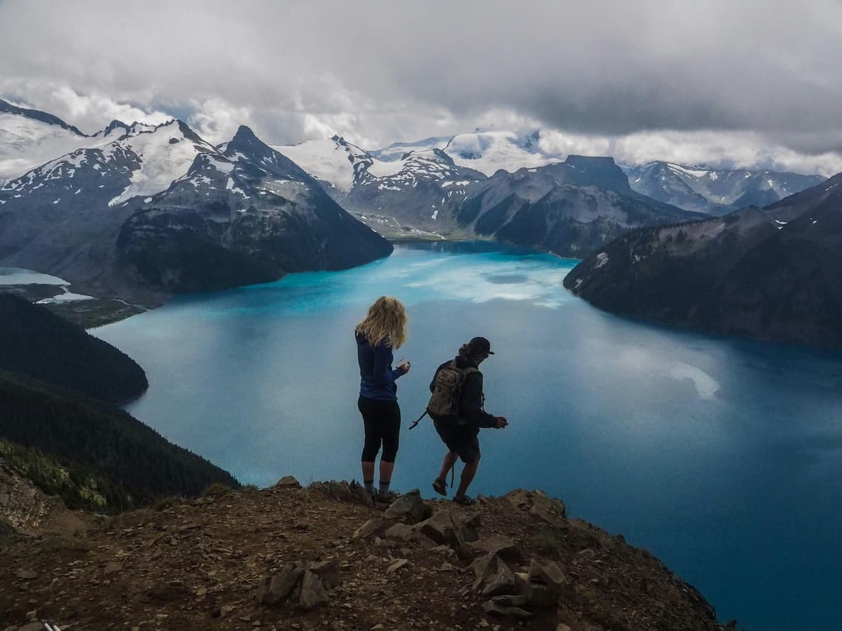 A man and a woman hiking on mountain with blue Garibaldi Lake and mountains in background