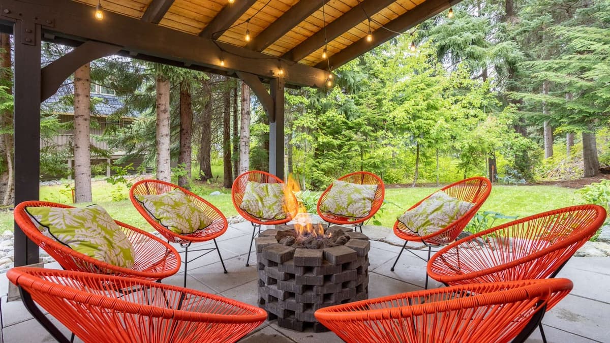 Outdoor patio with eight orange chairs surrounding a fire pit with lights on wood roof above looking out onto garden surrounded by tall green trees