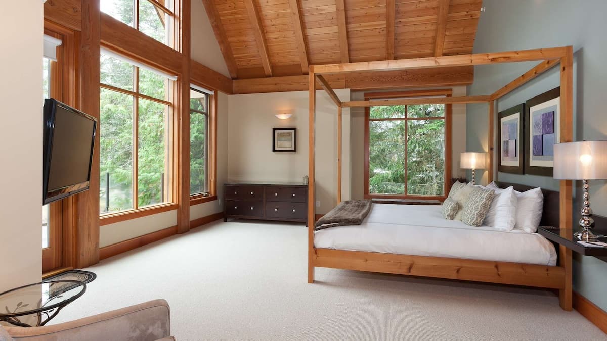 Master bedroom with cream carpets with wooden four poster bed and tall ceilings with multiple windows looking out onto tall green trees