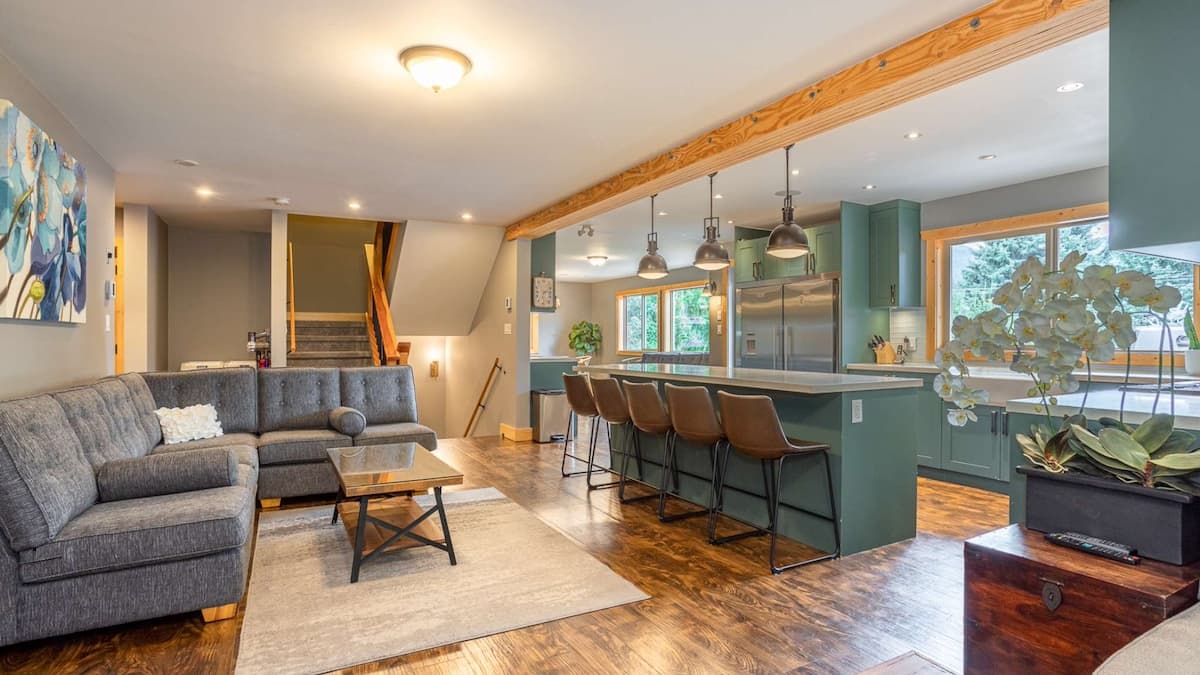 Integrated kitchen and living space with large grey L shaped couch and sage coloured kitchen with large kitchen island and 5 stools tucked into it