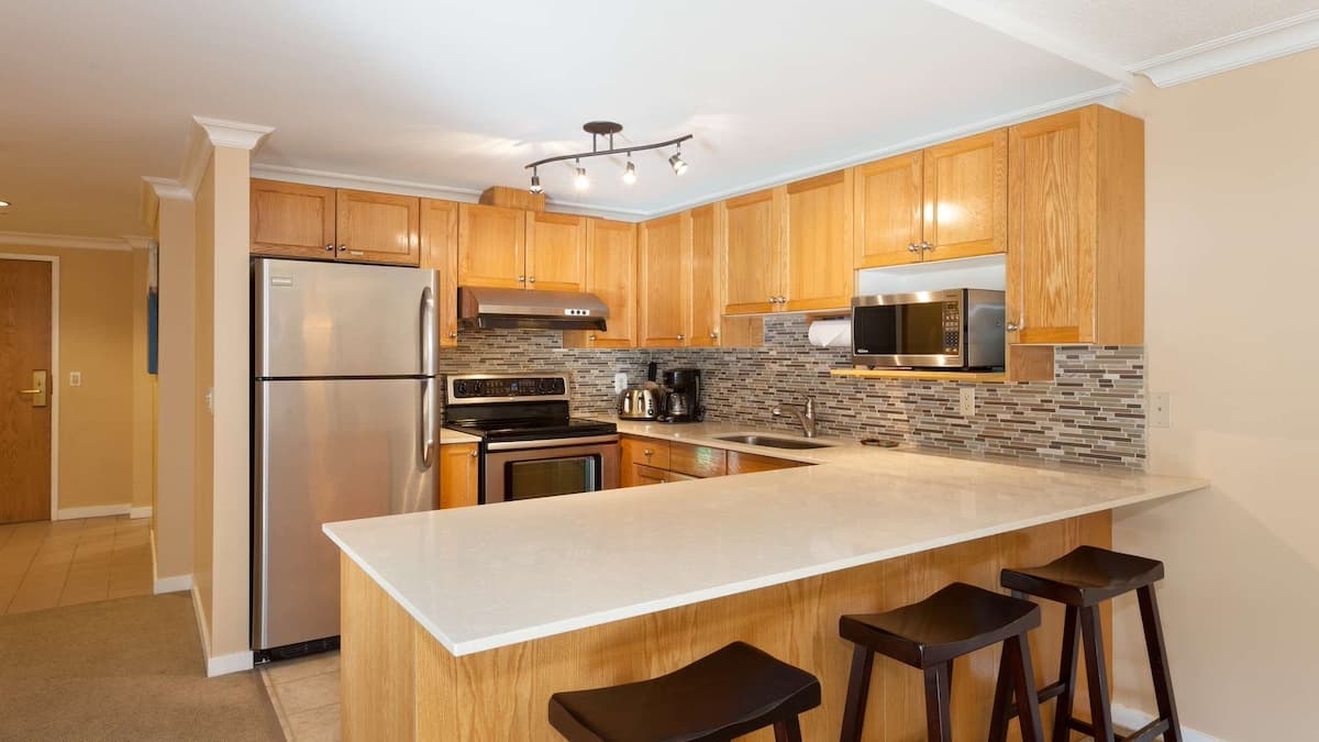 Large light brown wooded kitchen with modern appliances and large white kitchen counter with three black stools