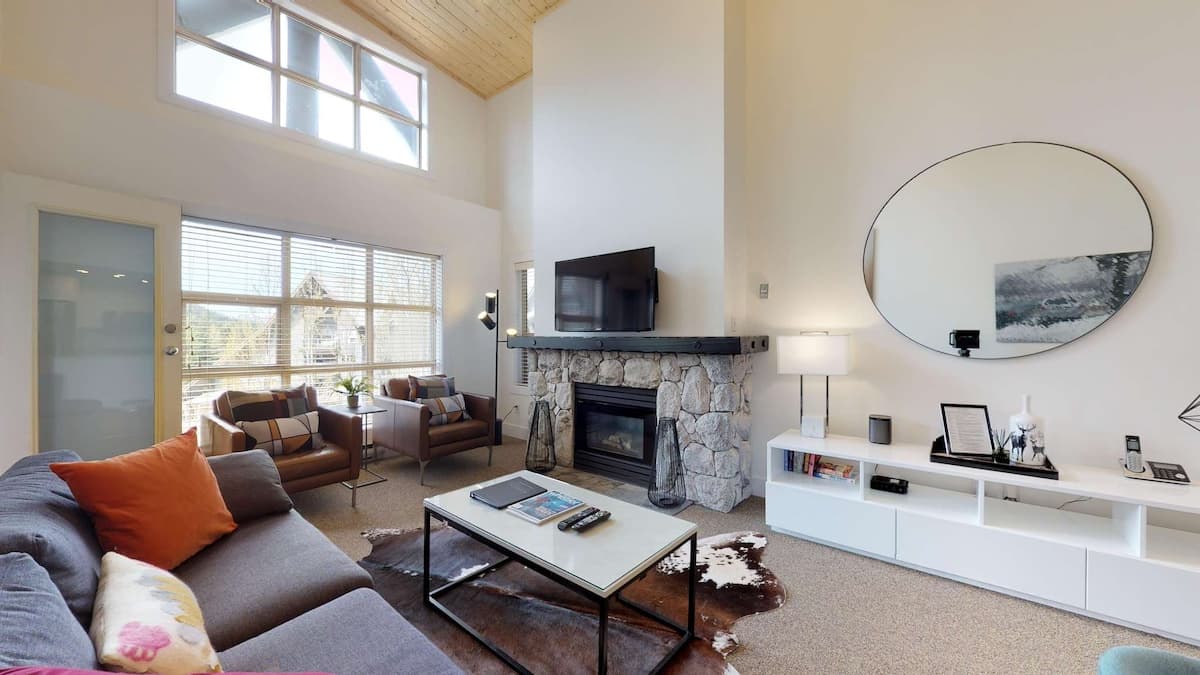 Living room with tall ceilings and big windows letting in natural light. Grey couch with two brown leather chair with white coffee table and large mirror and tv over the stone fireplace