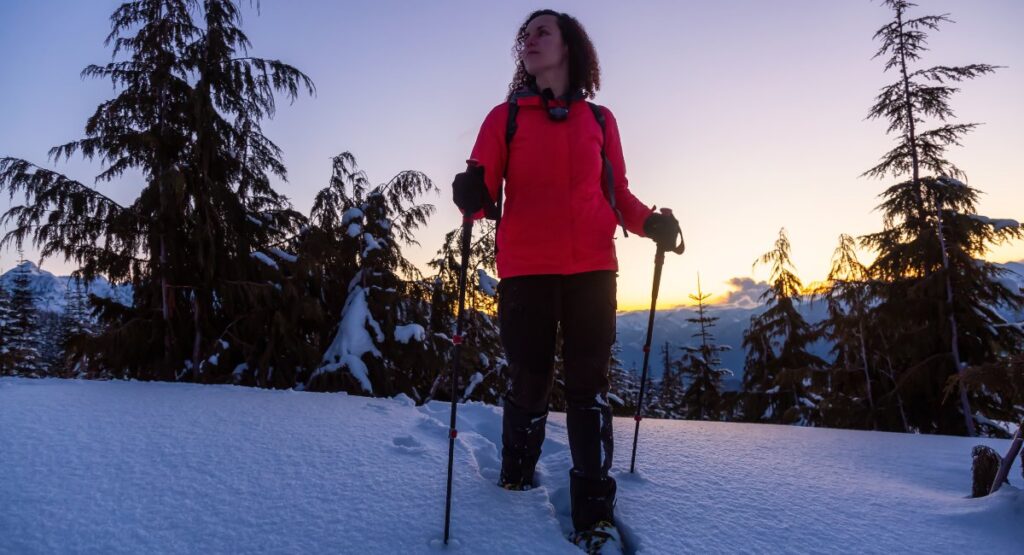 Adventurous woman snowshoeing in Whistler, with a picturesque view of snow-covered trees and distant mountain peaks illuminated by the setting sun.