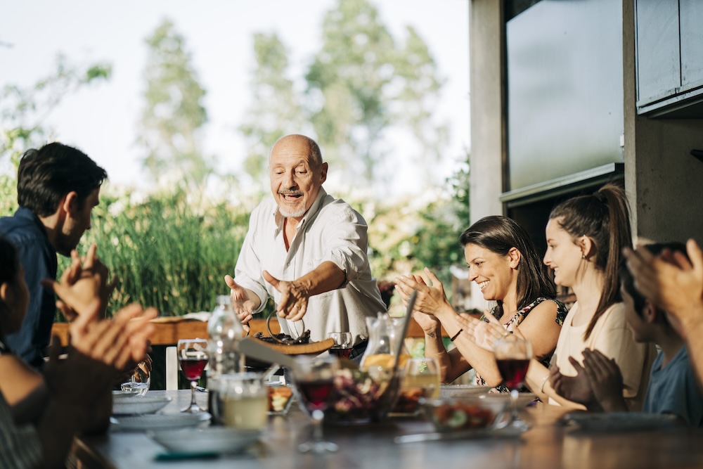 Cheerful senior man placing barbecue meat on tray over dining table for family. Multi-generational family at dining table in patio on a weekend.