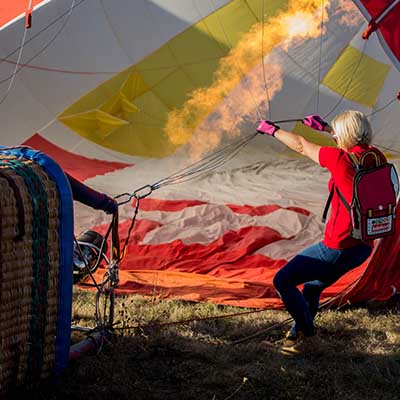 A person in a red jacket using a burner to inflate a hot air balloon on the ground.