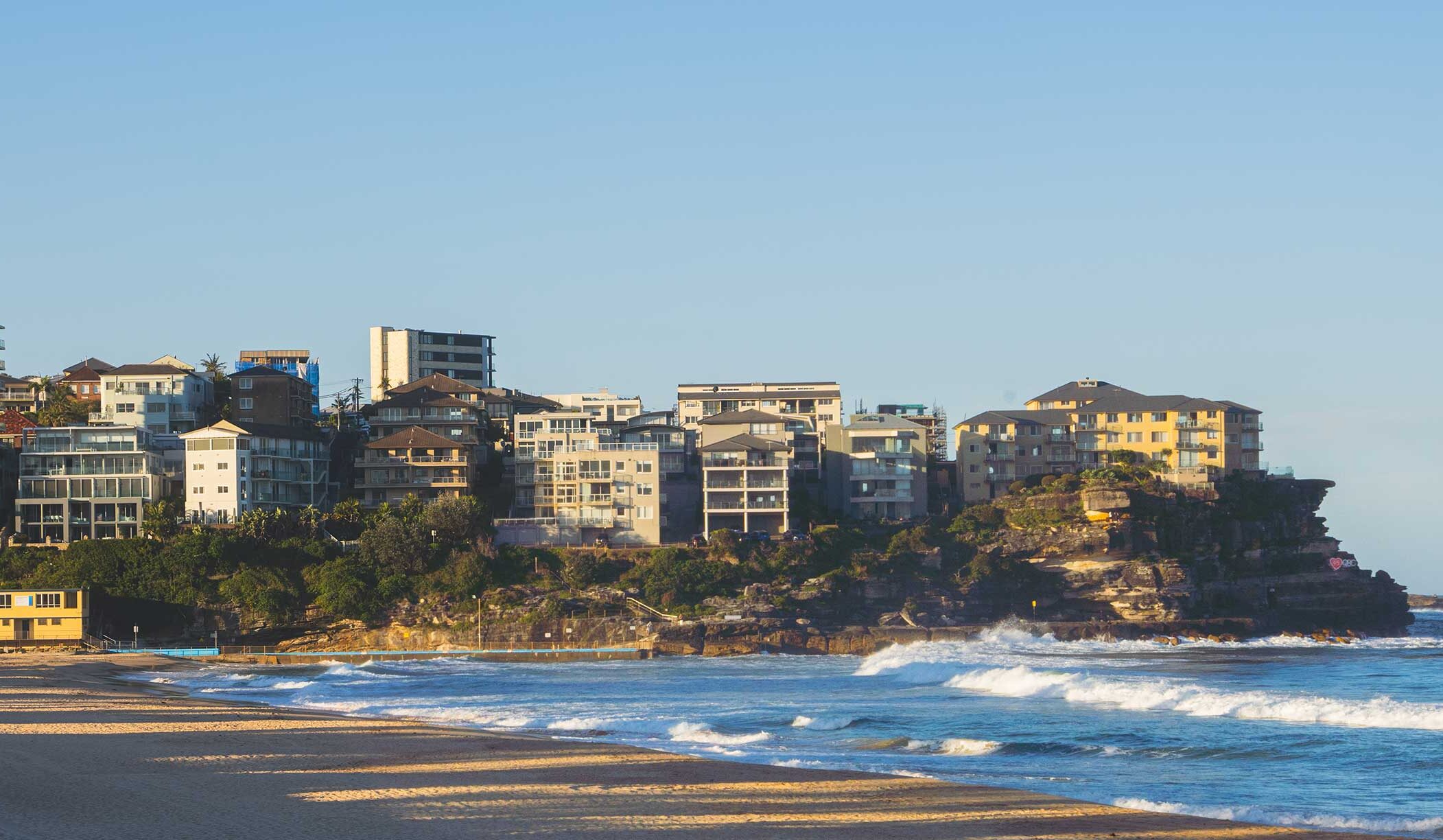 Modern luxury apartments overlooking Manly Beach in Sydney with gentle waves, under a clear sky at golden hour.