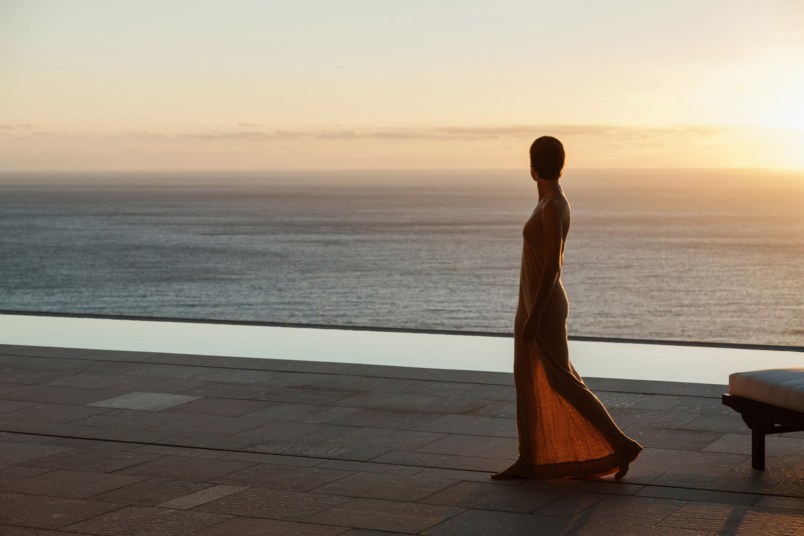Silhouette of a woman in an elegant dress standing by an infinity pool overlooking the sea at sunset.