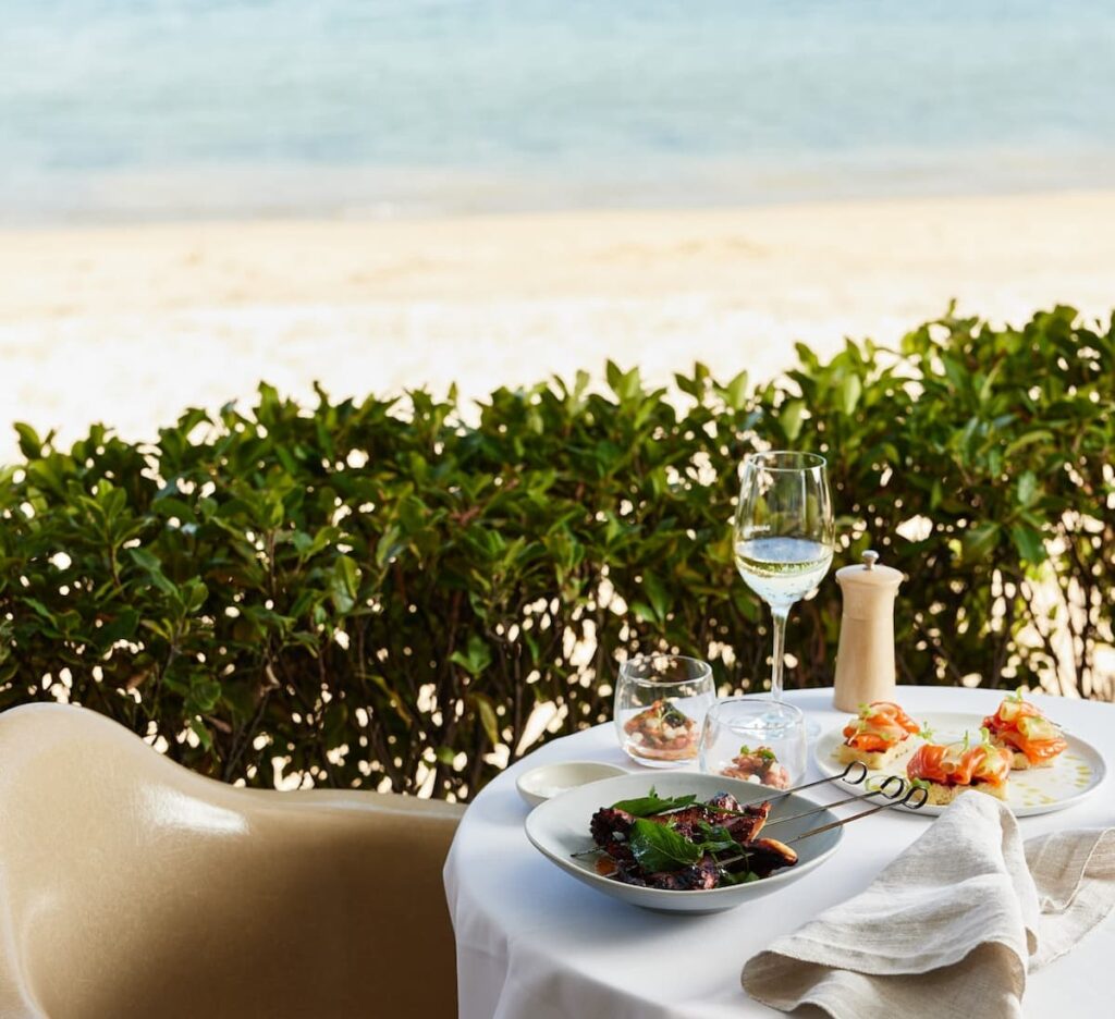 White linen table with plates of food and a wine glass beside a green hedge outdoors overlooking the beach