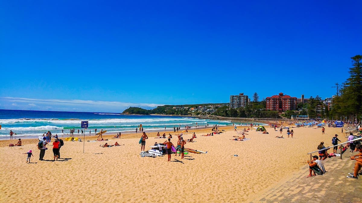 Crowds gathered on the sand at Manly Beach on a sunny day with clear blue skies