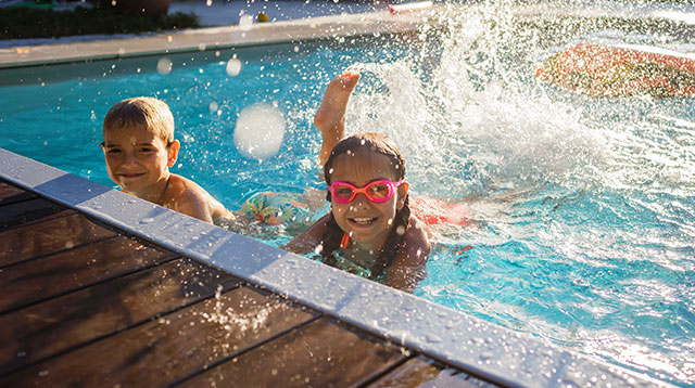 Two smiling children splashing water in an outdoor pool, enjoying a sunny day during school holidays.