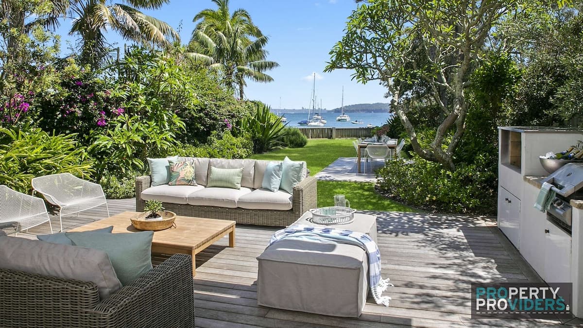 Outdoor patio with furniture in a private garden with lush greenery overlooking Little Manly Beach with sailboats in the water