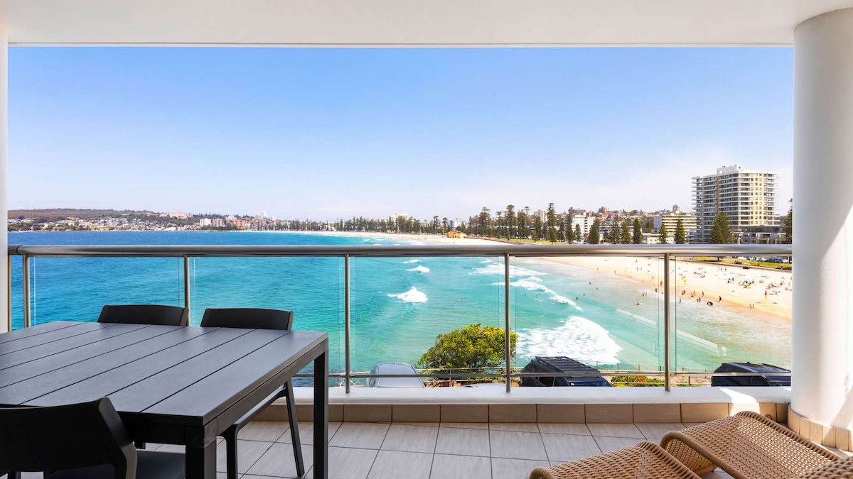 Spacious balcony with black table and chairs and two sun chairs overlooking Queenscliff pool and beach