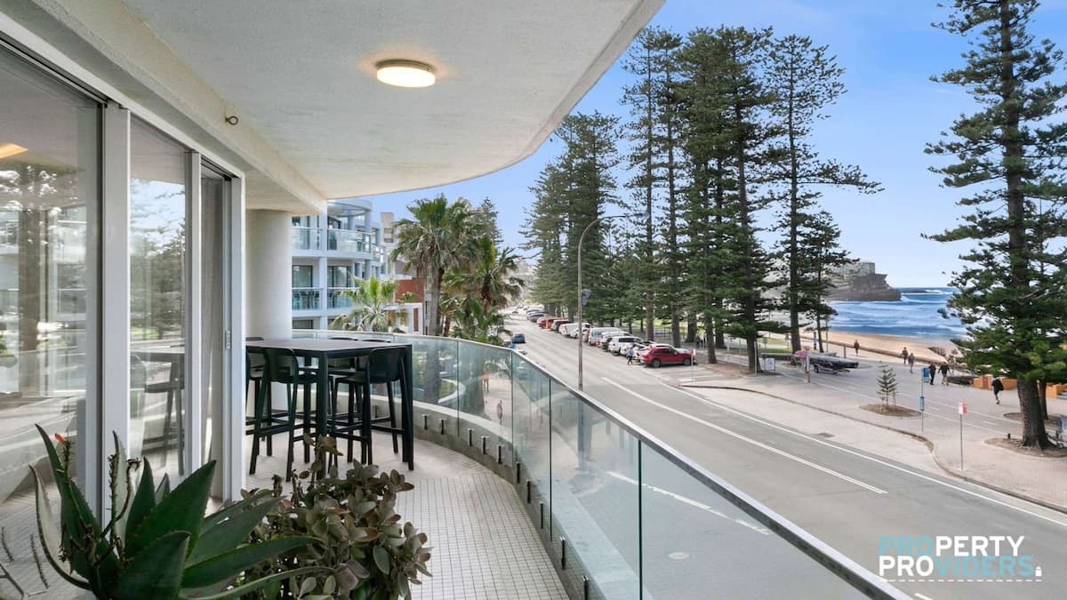 Balcony with green plants and black high chairs and table overlooking Manly Beach with tall green trees and blue ocean in background