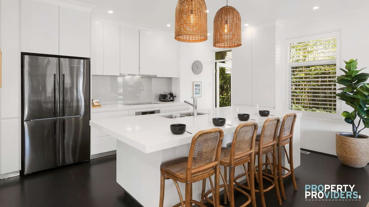 Large modern white kitchen with big island and 4 wicker stools. Large double door fridge to the left and large green houseplant on the right