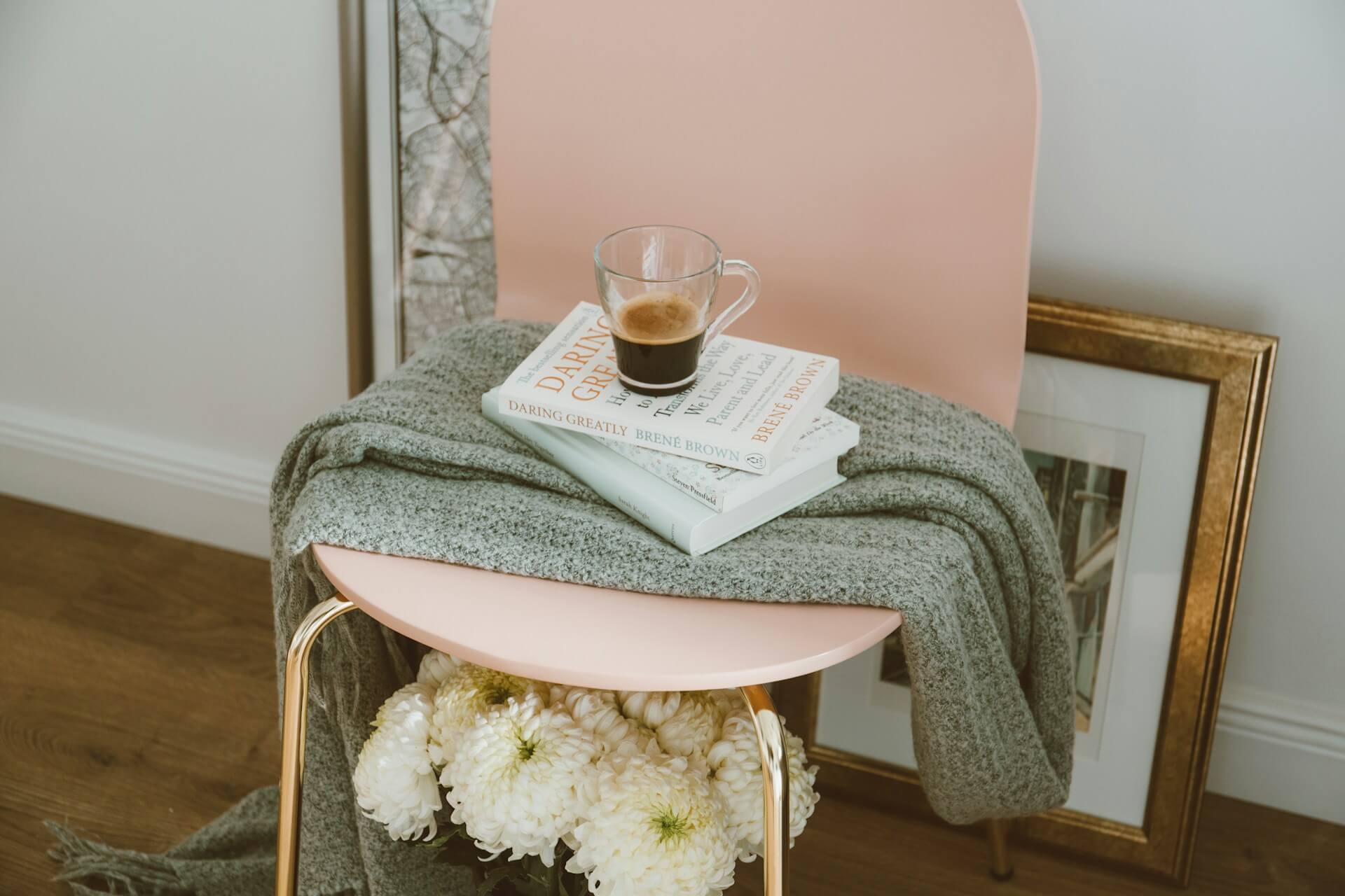 Cosy reading nook with a pink chair, a grey blanket, a stack of books, and a coffee cup. Fresh white flowers and a framed picture on the floor add to the serene atmosphere.