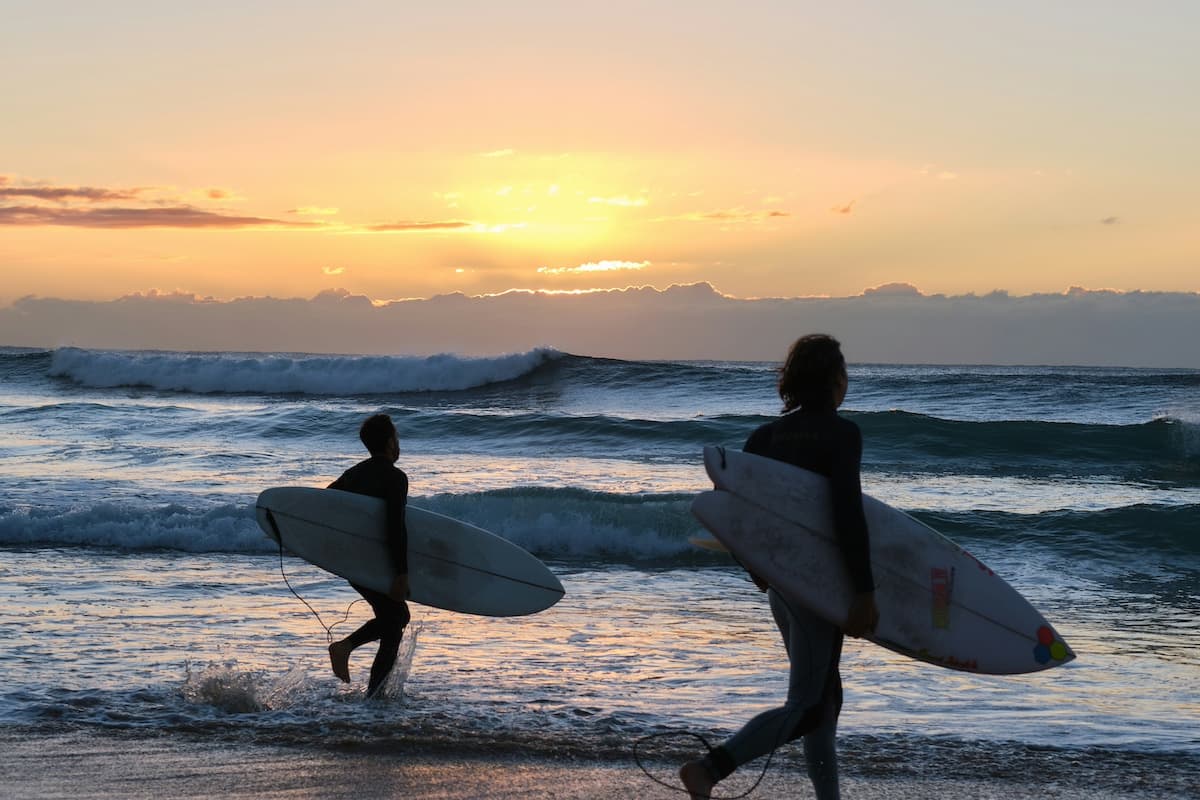 Two surfers carrying surfboards at Manly Beach with waves and sun setting in background