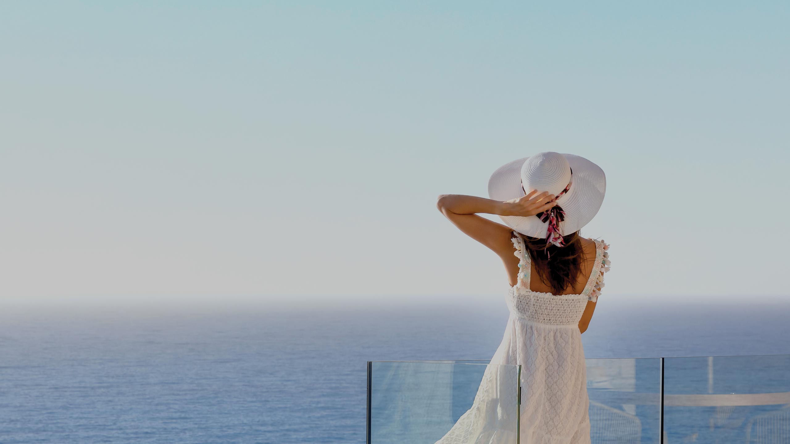 Woman in a white dress and wide-brimmed hat standing on a balcony overlooking the ocean, embodying peaceful luxury and serene vacation vibes.