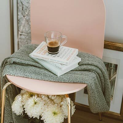 Cosy reading nook with a pink chair, a grey blanket, a stack of books, and a coffee cup. Fresh white flowers and a framed picture on the floor add to the serene atmosphere.