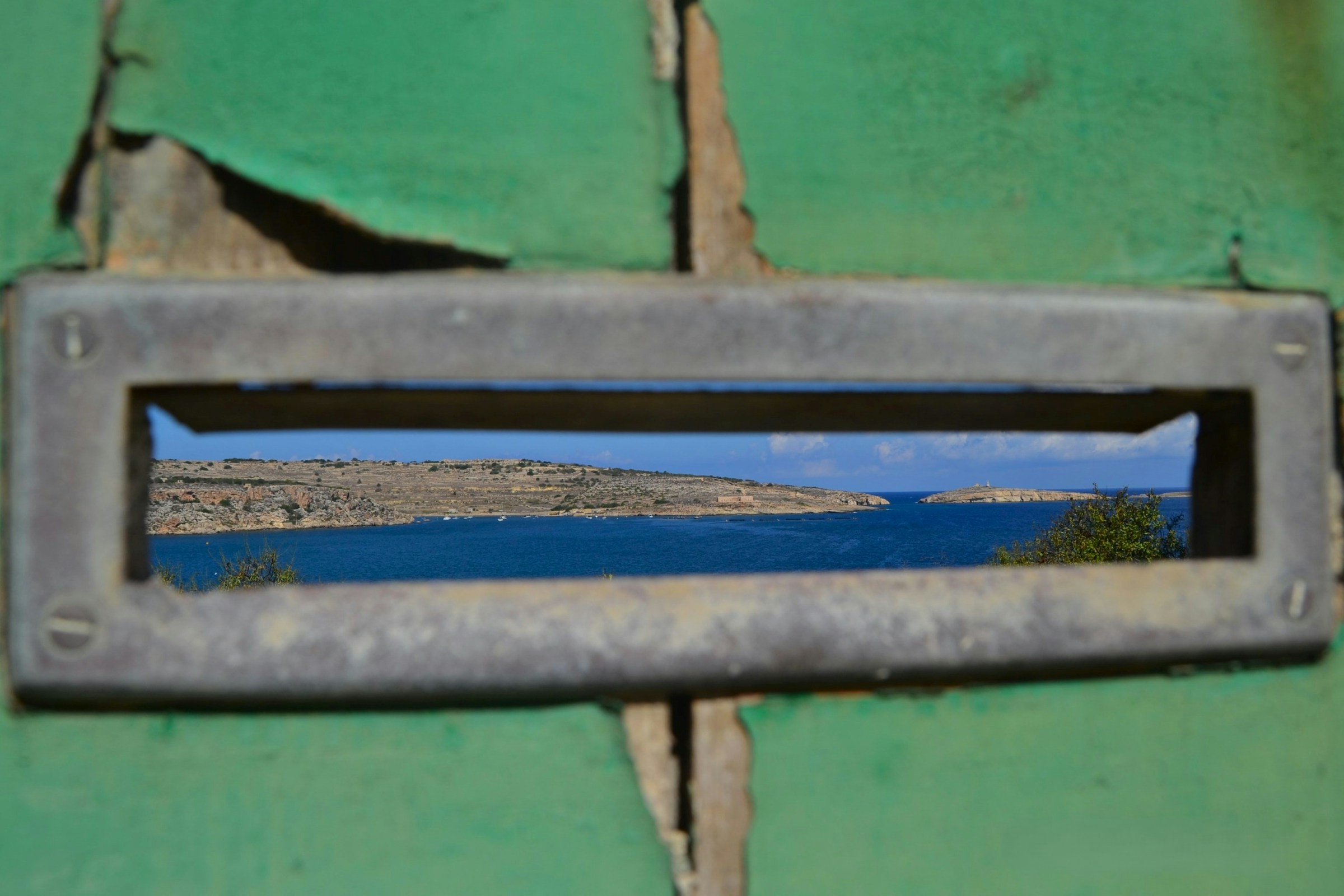 View through a mail slot in a weathered green door, overlooking a bright seascape, symbolizing the broader perspective needed in managing short-term rental cancellations.