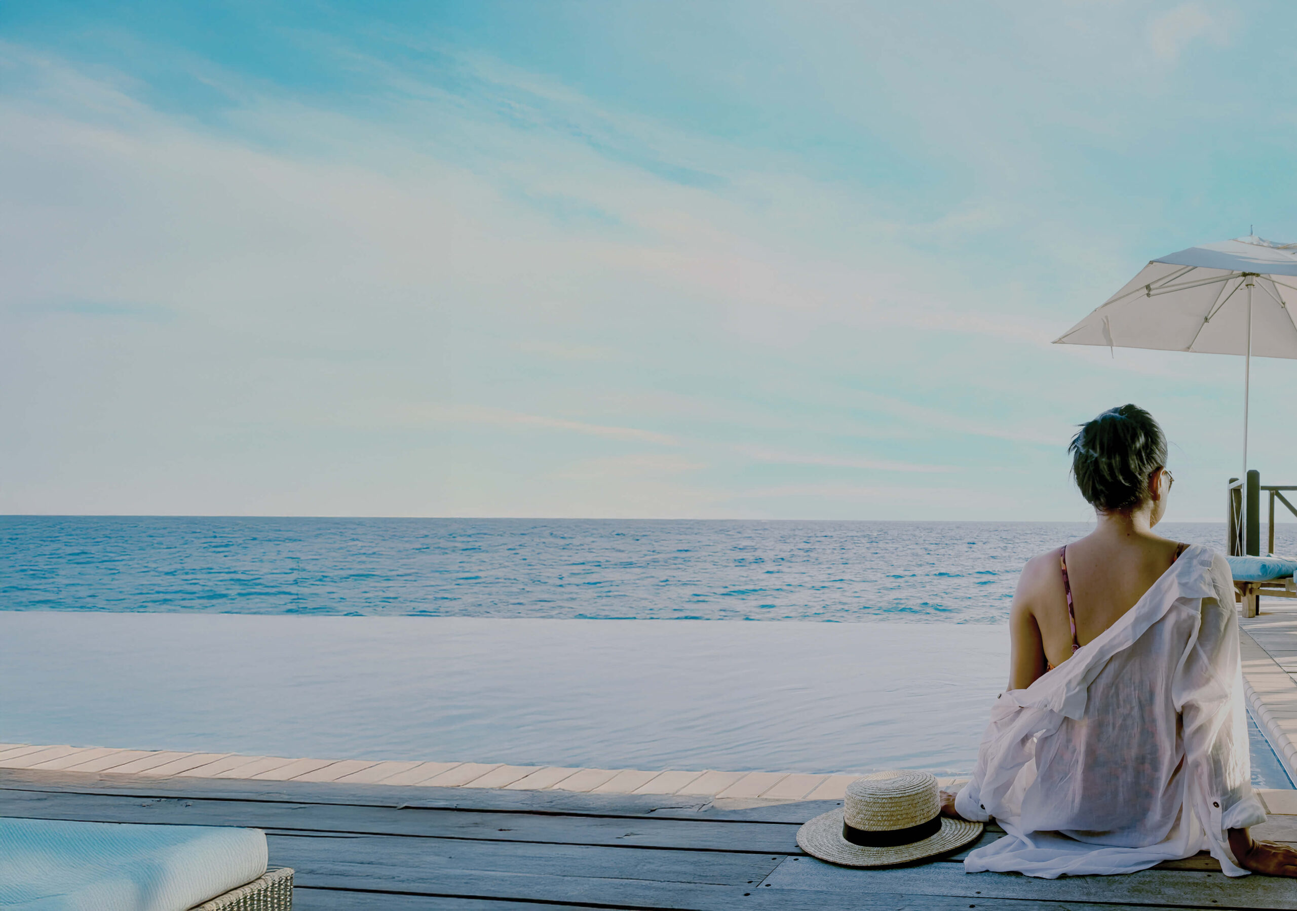 A woman relaxes by an infinity pool overlooking the ocean, dressed in a light cover-up, with a sunhat placed beside her. The scene exudes tranquillity, luxury, and coastal serenity.