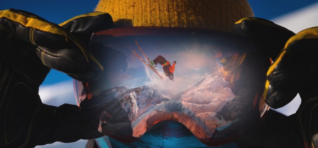 skier in goggles, in which we see the reflection of another skier doing a backflip on a slope