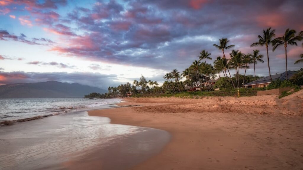 Kamaole Beach with green palm trees lining the shore at sunset with mountain in the background