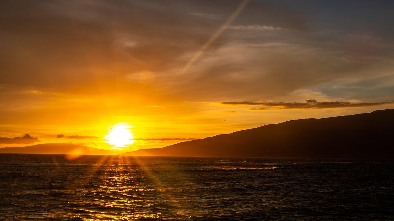 Brilliant Maui sunset with the sky painted in hues of orange, reflecting over the calm ocean waters, with a silhouette of Maui and distant islands on the horizon