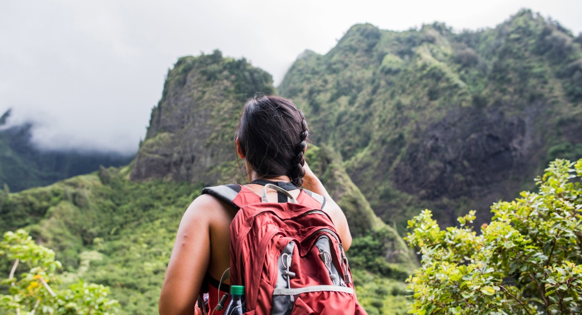 Solo hiker with red backpack admiring lush green peaks and misty cliffs in ʻĪao Valley, Maui