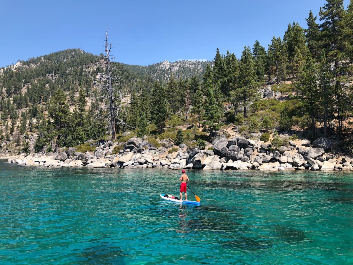 Man on paddleboard on Lake Tahoe with rocks and trees on the shore