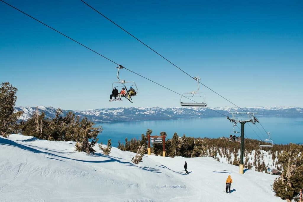 People skiing on Heavenly Mountain with people sitting on chair lift and blue waters from lake tahoe in background