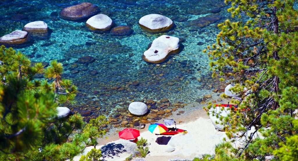Crystal-clear waters of Lake Tahoe with smooth white stones and colorful beach umbrellas on a sandy shore, surrounded by vibrant green pine trees, a serene vacation spot.