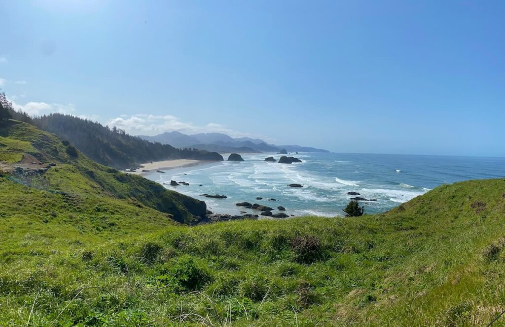 View from a hiking trail in Ecola State Park surrounded by green grass and a view of the coast with rock formations in the water