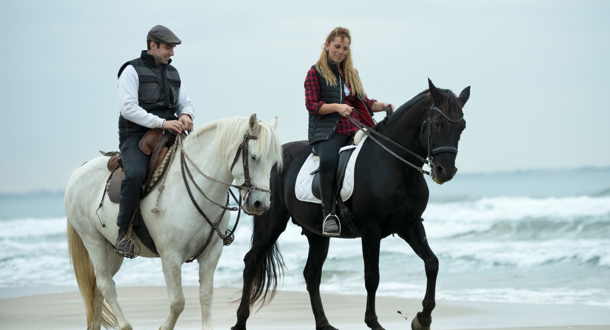 Two riders on horseback at the beach, one on a white horse and the other on a black horse