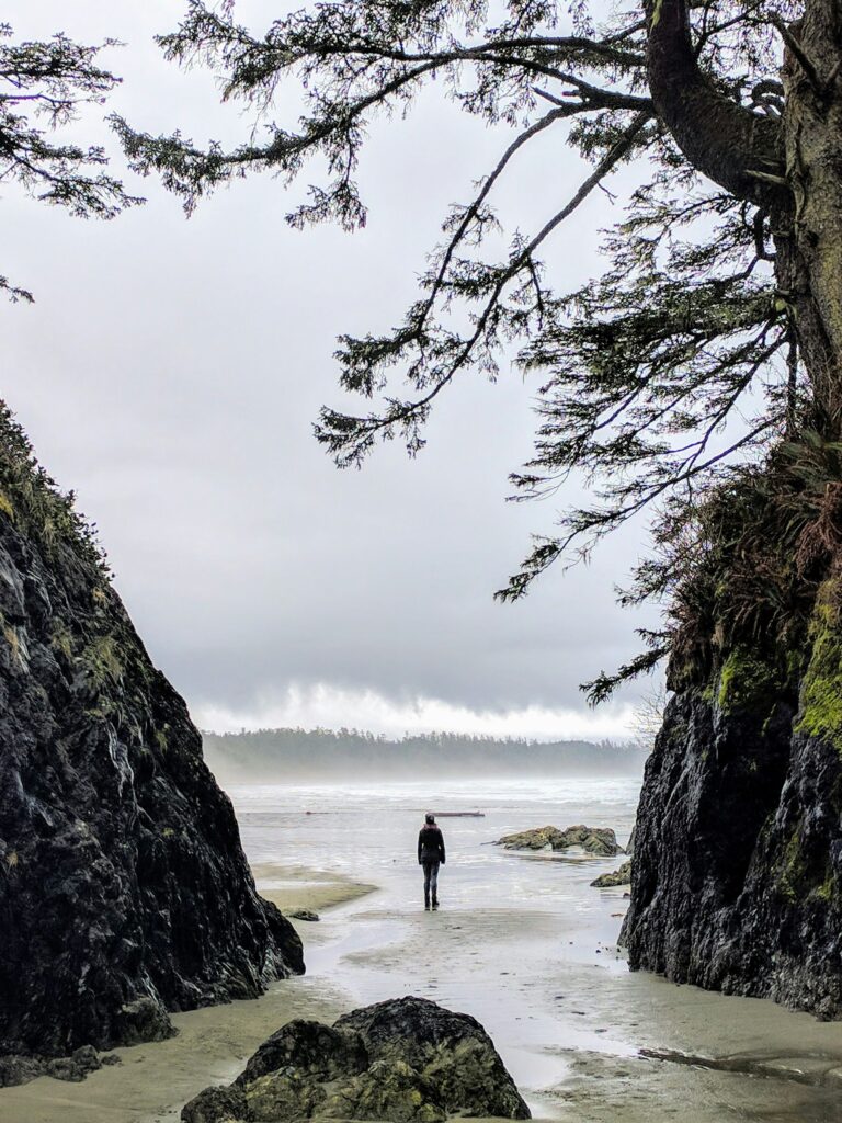 person standing between rock formations on a beach in the winter with a large wave crashing on the beach 